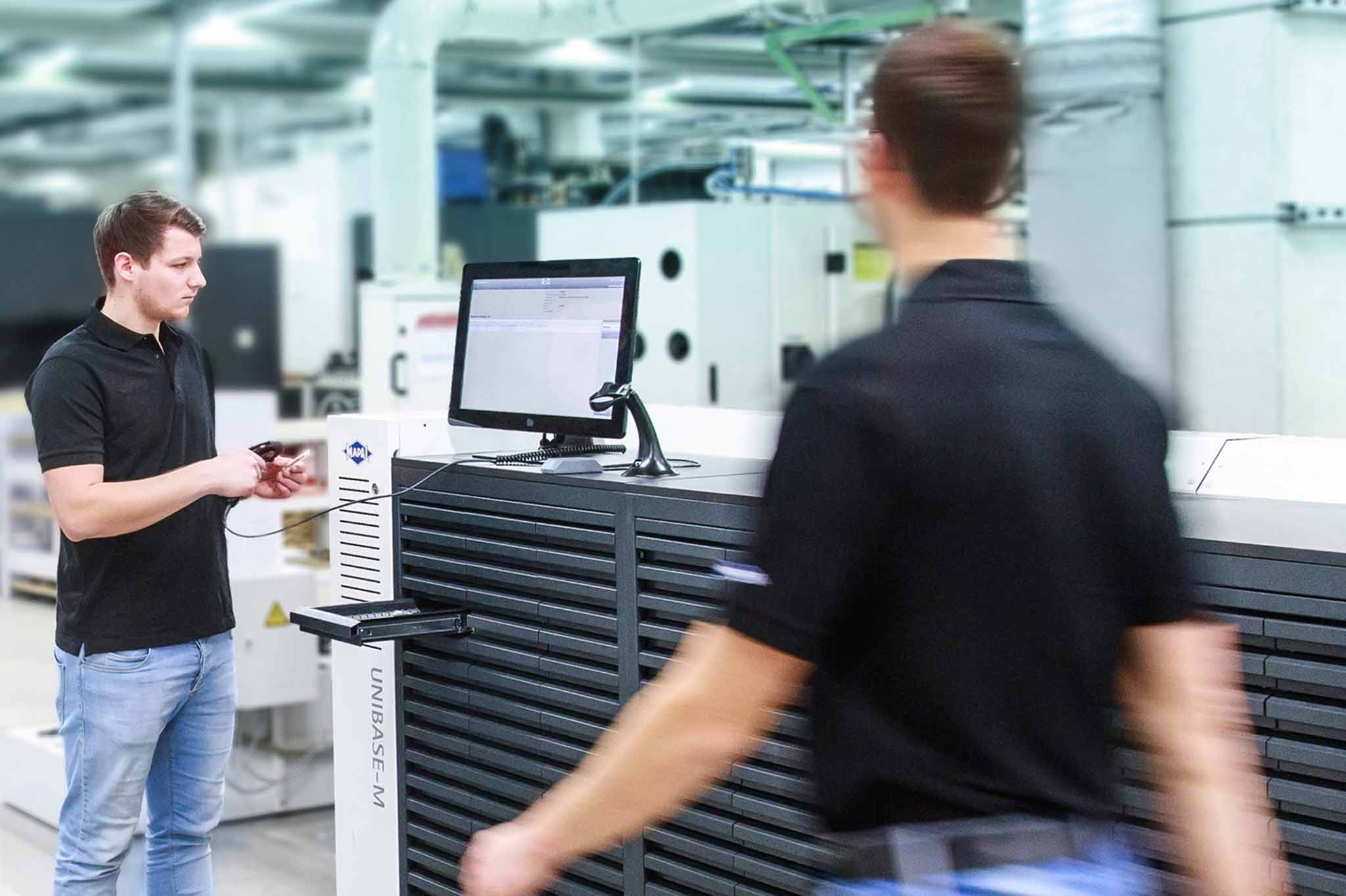 Ein Mitarbeiter steht vor einem Werkzeugausgabesystem UNIBASE-M. Ein Mitarbeiter läuft vorbei.